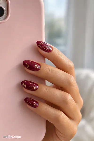 Close-up of almond nails with burgundy polish and delicate gold foil flakes