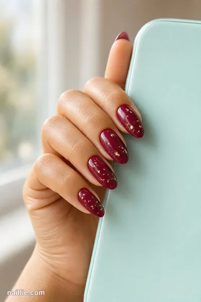 Close-up of almond nails with burgundy polish and delicate gold foil flakes — look 2