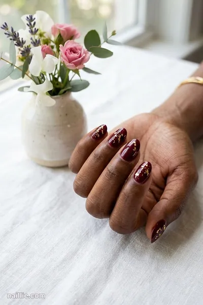 Close-up of short almond nails with burgundy polish and delicate gold foil flakes