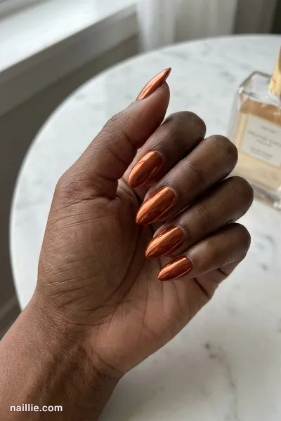 Close-up of almond nails with burnt orange polish and a mirror-like chrome finish