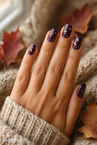 Close-up of short square nails with deep plum polish and delicate gold foil flakes
